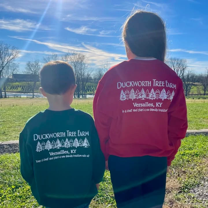 Two children wearing Duckworth Tree Farm shirts stand with their backs to the camera, with the farm’s tree rows beyond.