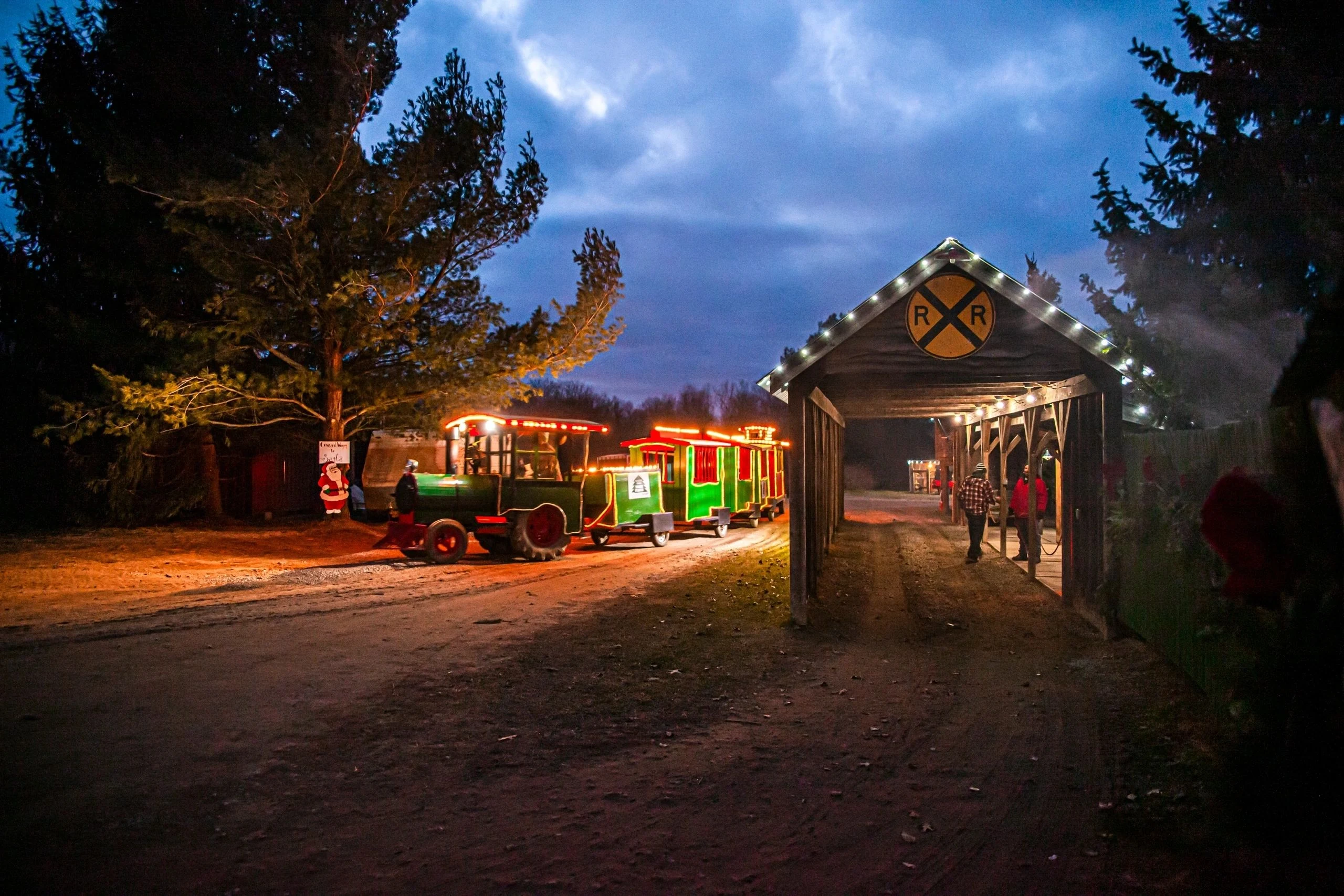 A decorated green train and the lit station at Peacock Road Family Farm at dusk.
