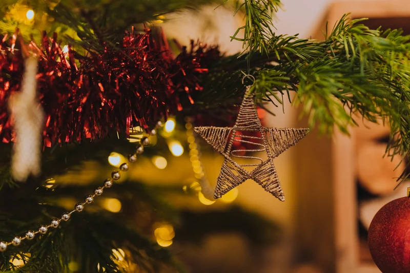 A gold openwork star hangs among natural evergreen branches, with red garland, a gold bead strand and warm lights behind it.