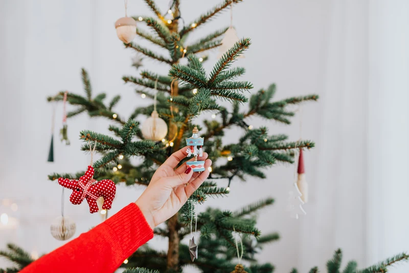 A hand placing a small carousel ornament on a real Christmas tree decorated with wooden and fabric ornaments.