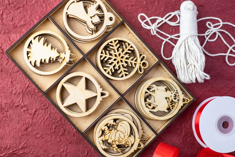 A compartmented box of wooden stars, snowflakes and other Christmas ornaments beside hanging cord and ribbon on a red surface.