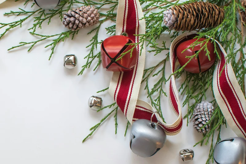 Red-and-cream striped ribbon lies in loose loops beside red and silver bells, pinecones and evergreen sprigs on a pale surface.
