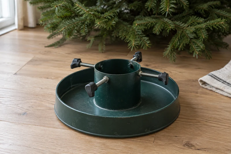 A green Christmas tree stand with four screw clamps and a broad reservoir on a wooden floor.