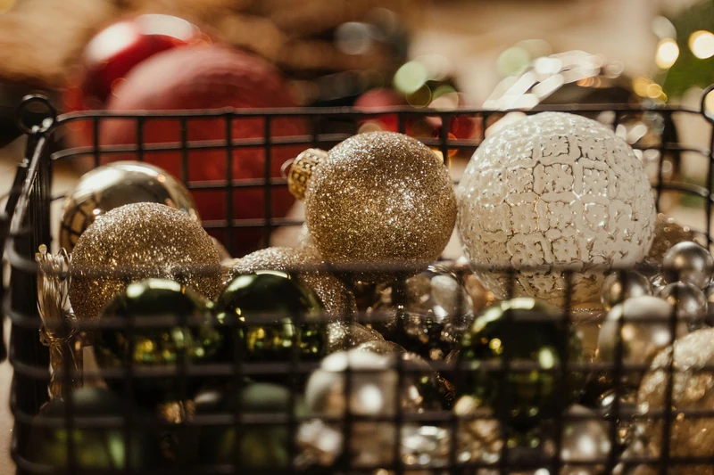 Gold, silver and green baubles gathered in a black wire basket, with softly blurred Christmas lights behind them.