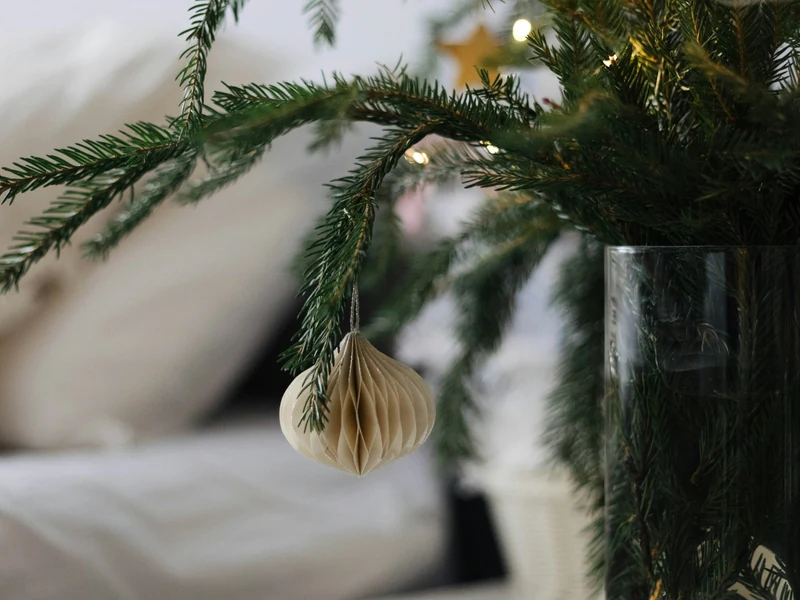 A cream honeycomb paper ornament hanging from cut evergreen branches arranged in a clear glass vase.